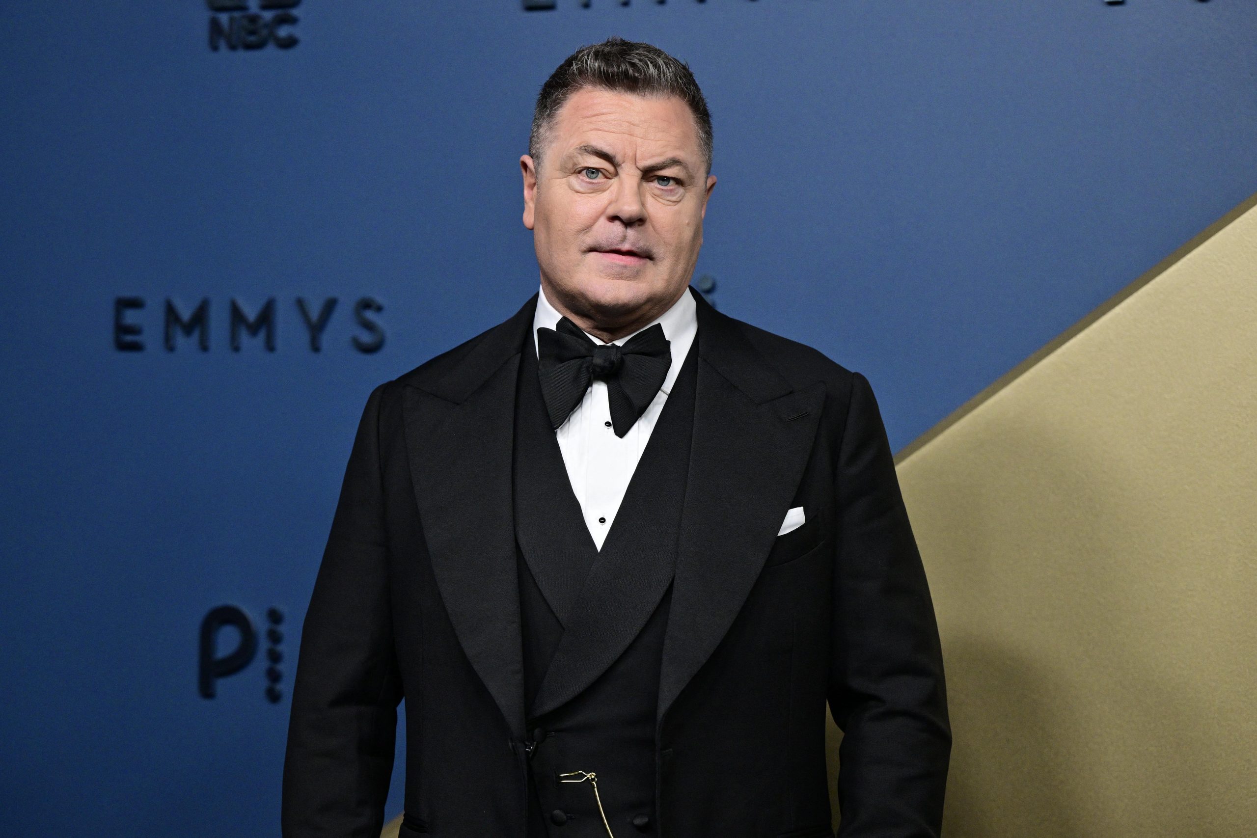 Nick Offerman wearing a black tuxedo with a bow tie and pocket square, standing in front of an Emmy Awards backdrop.