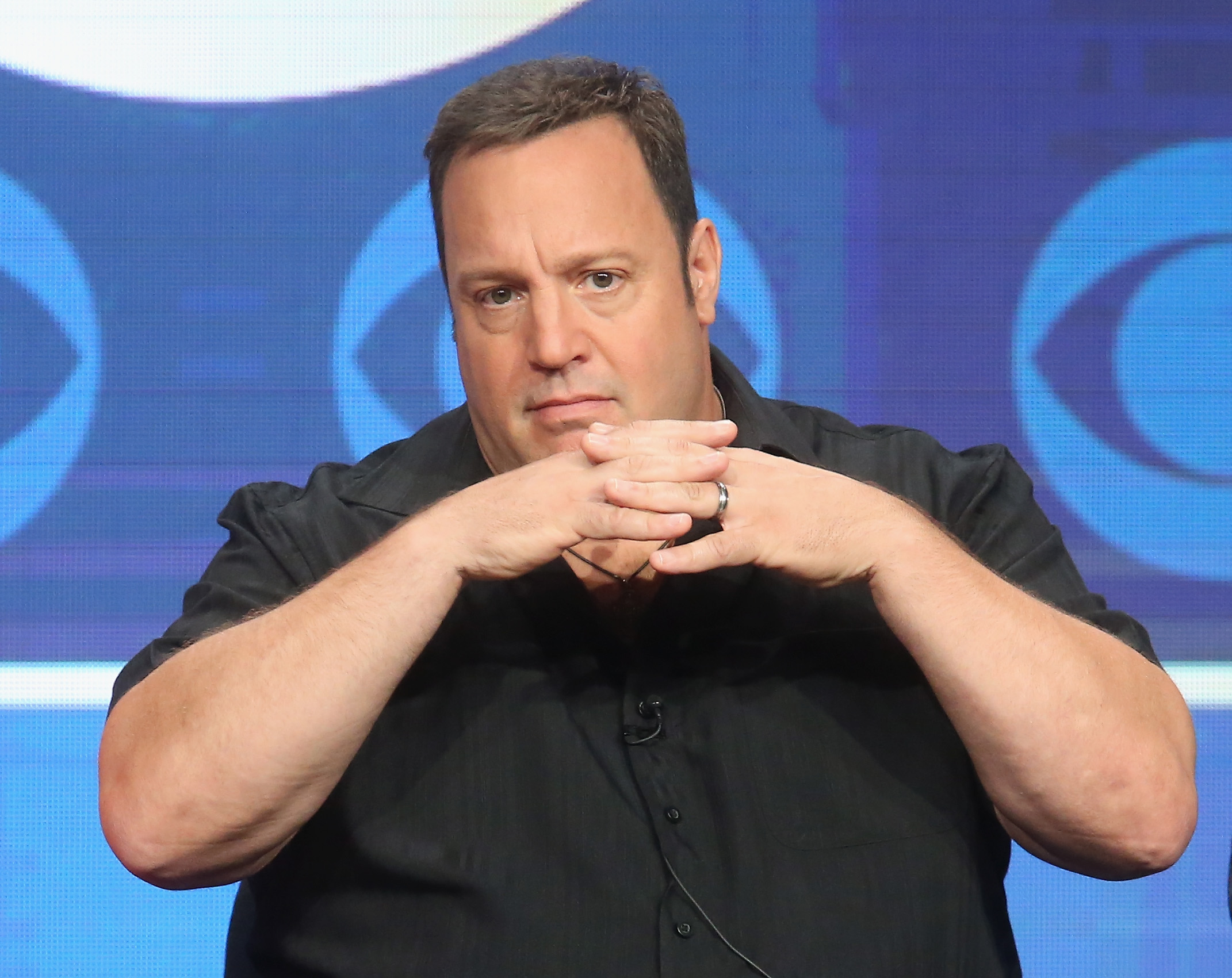 Kevin James in a black button-up shirt sits with his hands clasped in front of his face against a blue background.