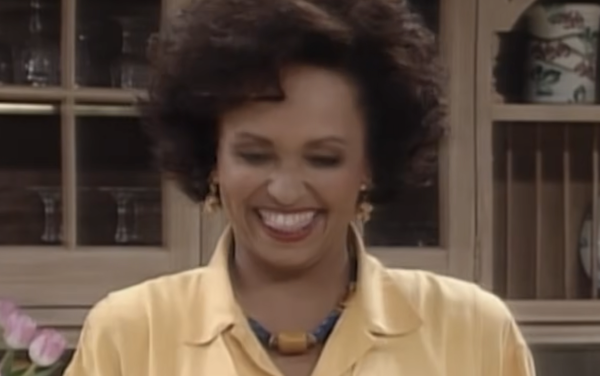 A woman with dark hair styled in a bouffant laughs while wearing a yellow collared shirt and a chunky statement necklace.
