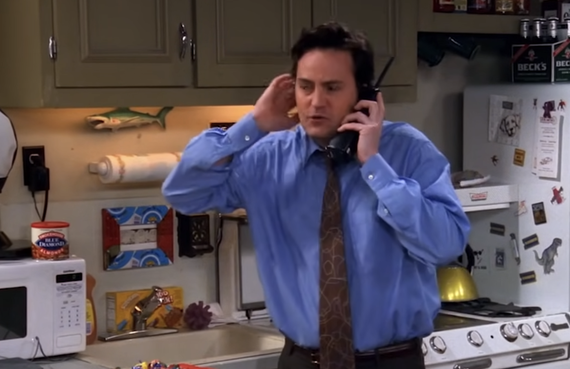 Chandler Bing in a blue dress shirt and patterned tie holds a black cordless phone to his ear while standing in a kitchen.