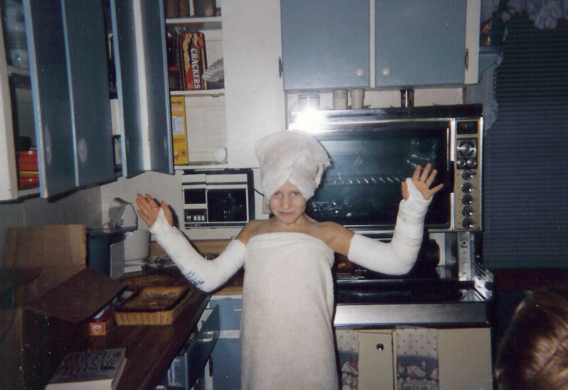 A child wrapped in a bath towel with a towel on their head, posing with both arms in white casts in a kitchen.