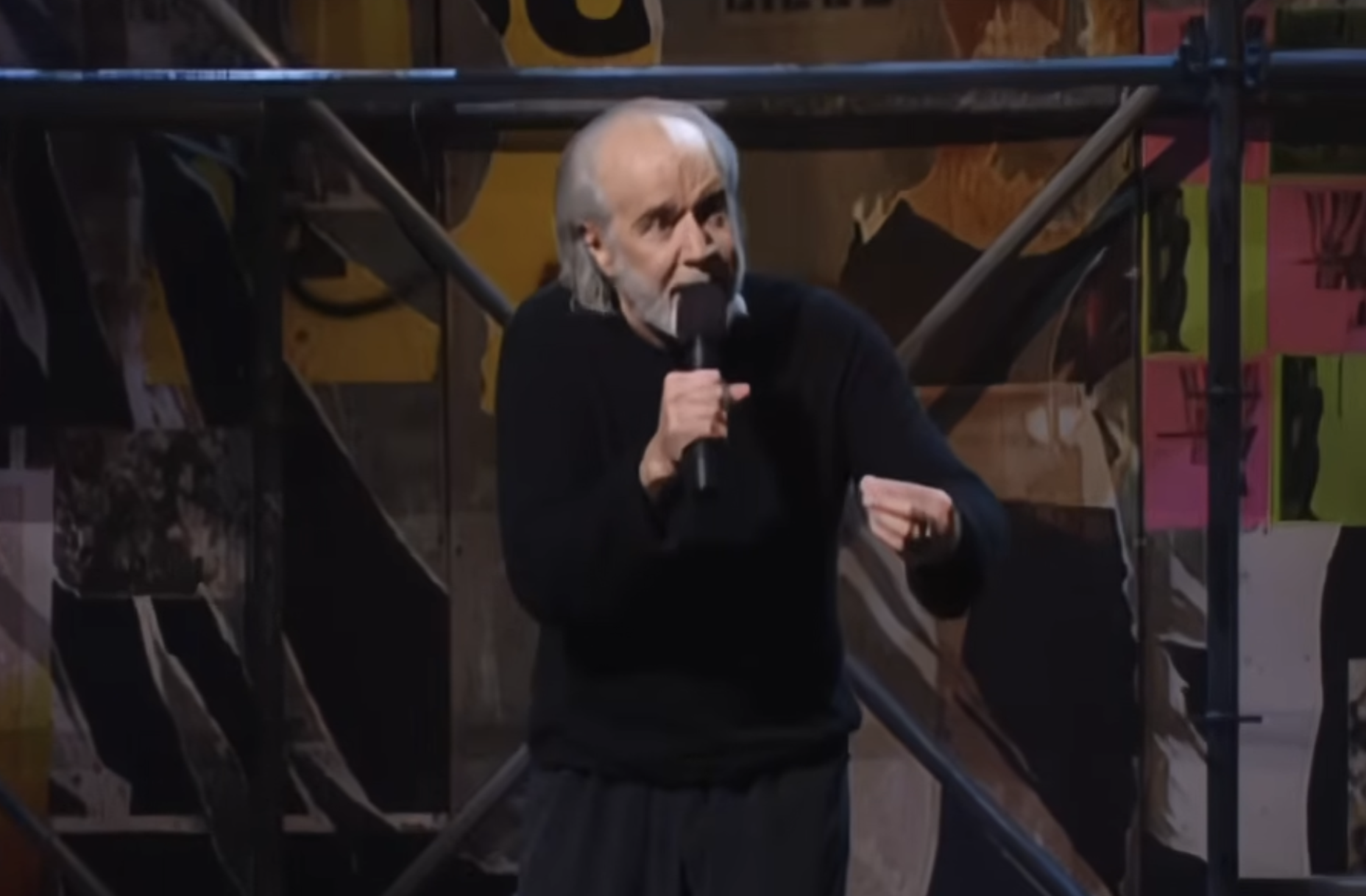 George Carlin performing stand-up comedy with a microphone on stage.