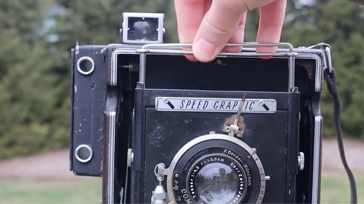 A hand adjusts the metal frame viewfinder on a vintage Speed Graphic folding camera.