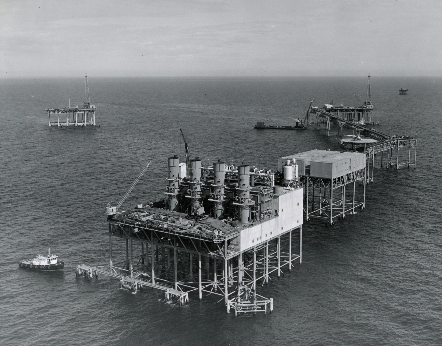 An aerial view of an offshore oil platform with multiple structures connected by walkways, including a main facility with large vertical tanks and a supply vessel nearby.