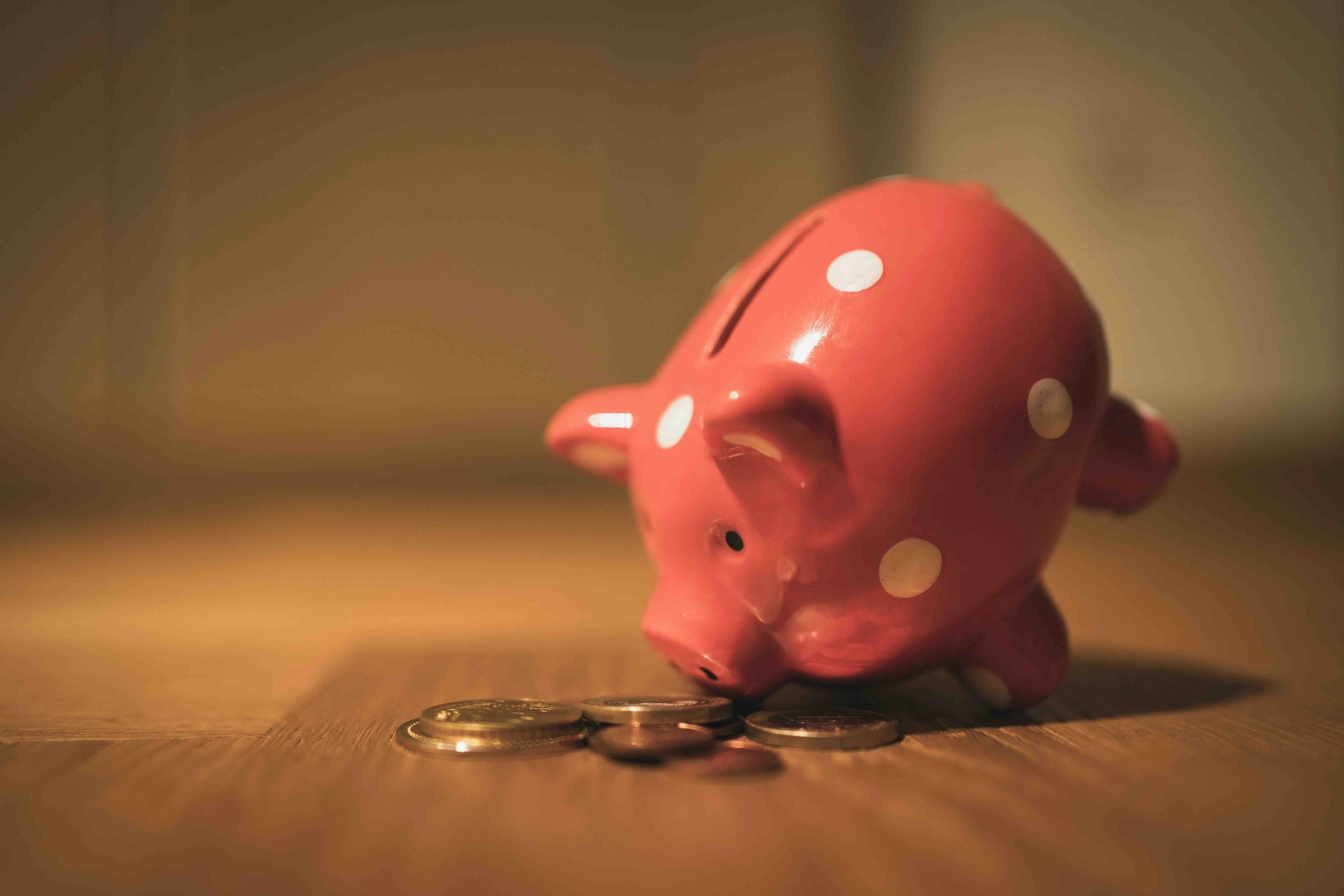 A pink piggy bank with white polka dots tipped over on a wooden surface next to a pile of coins.