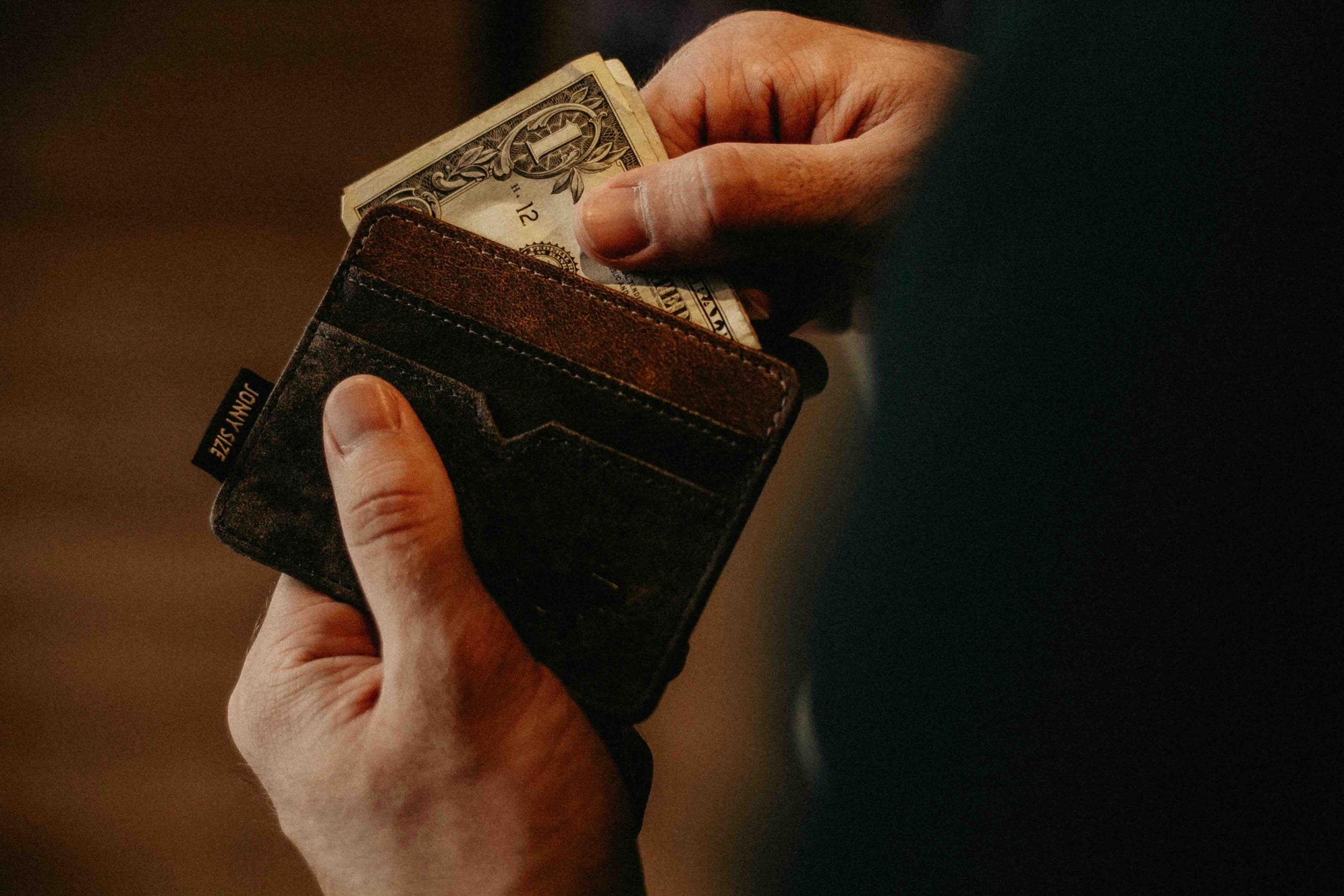 Hands sliding a one-dollar bill into a slim wallet labeled “JONNY SIZE.”