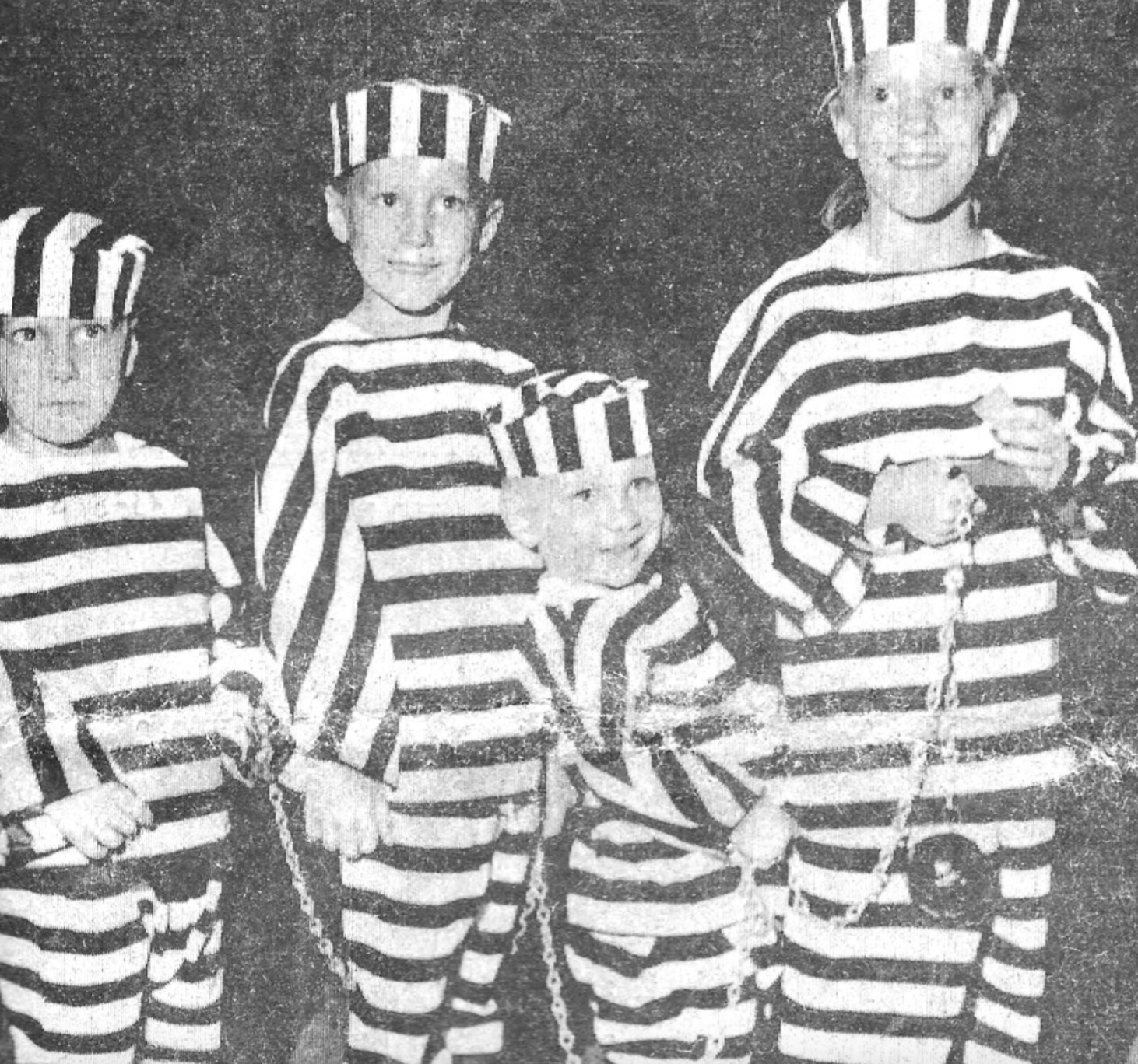 Four children wearing black-and-white striped prison uniforms and matching caps, connected by chains.