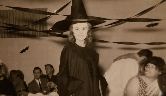 A woman in a pointed witch hat stands in a room decorated with paper bats and streamers at a party.