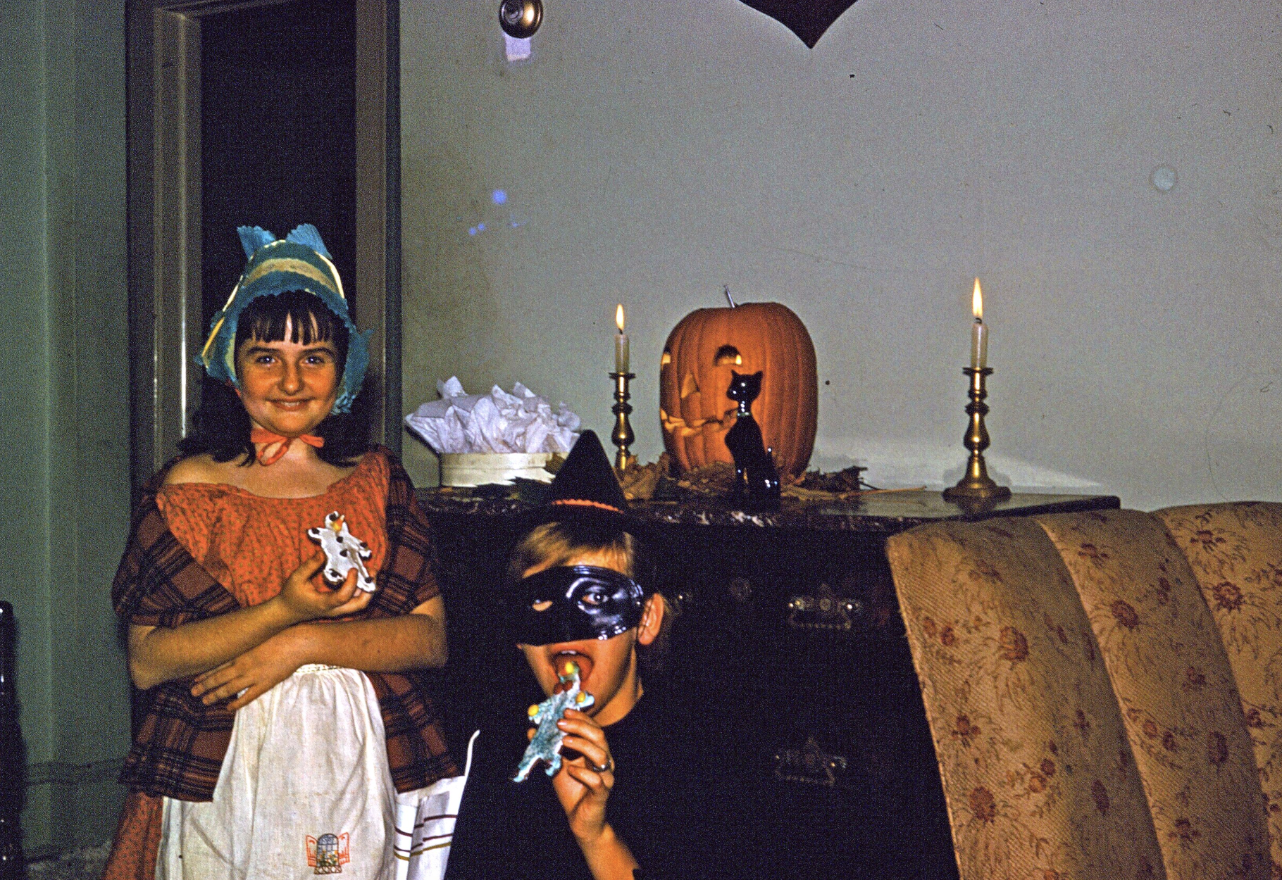 Two children in Halloween costumes pose in front of a carved pumpkin and two lit candles.