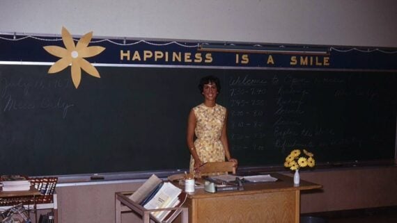 A teacher stands at her desk in front of a blackboard that reads “HAPPINESS IS A SMILE” and displays an open house schedule dated July 11, 1967.