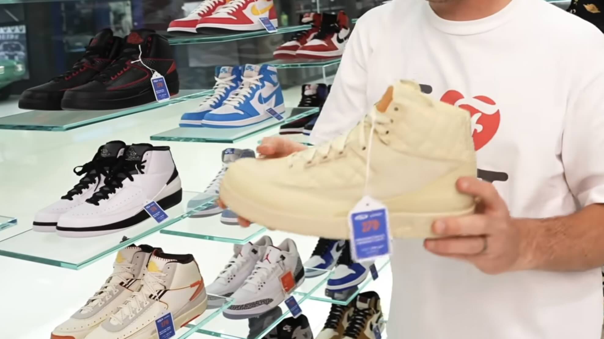 A person in a white t-shirt holding a beige high-top sneaker in front of store display shelves stocked with various athletic shoes.