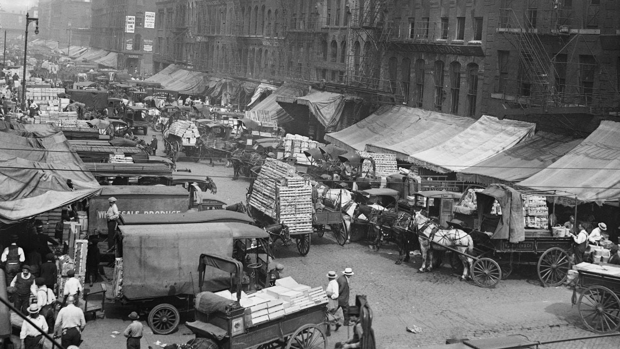 A busy city street in the early 1900s with horse-drawn carts, delivery trucks, market stalls, and people wearing hats and old-fashioned clothes. Large buildings and awnings line both sides of the street.