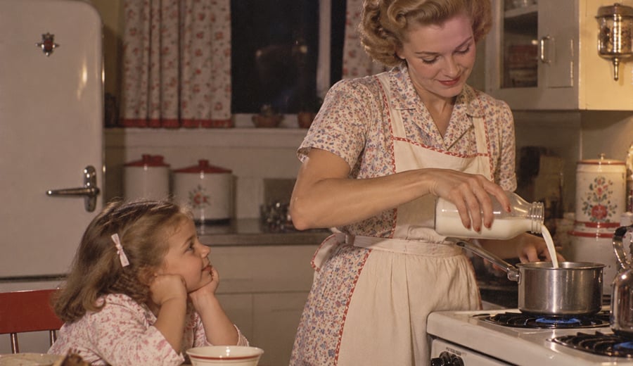 A woman in a vintage kitchen pours milk into a pot on the stove while a young girl sits at the table, watching her with a smile.