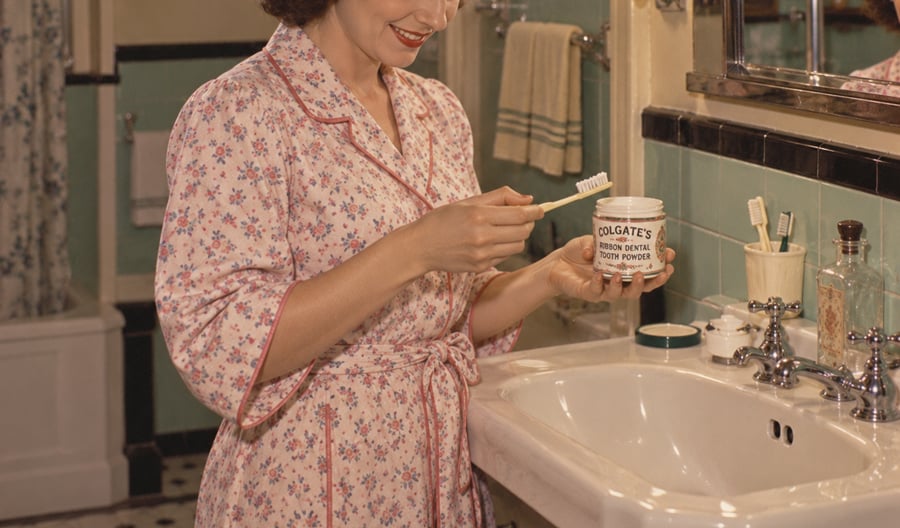 A woman in a floral robe stands at a bathroom sink, smiling as she applies Colgate’s tooth powder to a toothbrush. The bathroom has green tiles, a towel rack, and various bottles on the sink.