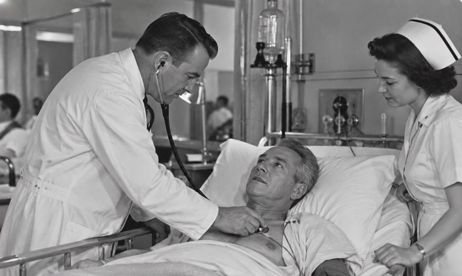 A doctor listens to a male patient’s chest with a stethoscope as a nurse stands by the hospital bed, both observing the patient. The scene appears to be from an older era, based on their uniforms and the equipment.