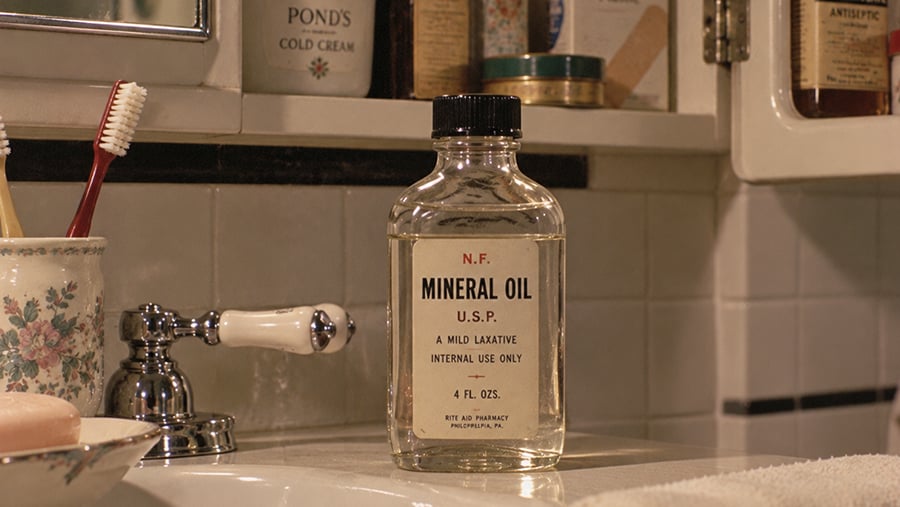 A clear bottle labeled “Mineral Oil” sits on a bathroom sink next to a floral cup holding toothbrushes, with toiletries and a bar of soap visible in the background.