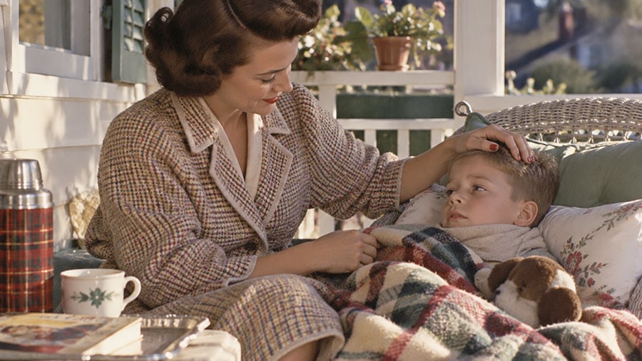 A woman sits on a porch comforting a young boy wrapped in a blanket, gently touching his head. A stuffed animal, a thermos, a book, and a cup are nearby, creating a cozy atmosphere.