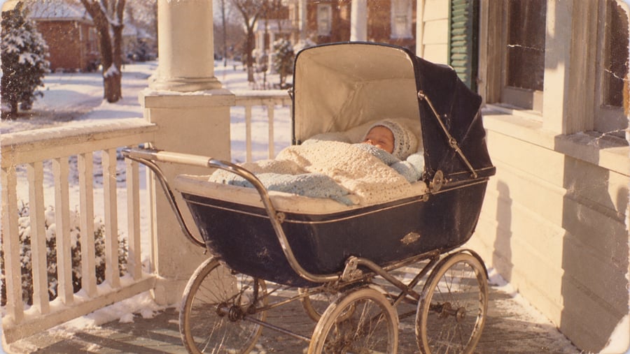A baby bundled in blankets lies in a vintage navy stroller on a snowy porch, with houses and trees visible in the background.