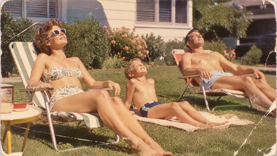 A woman, a child, and a man in swimsuits relax on lawn chairs in the sun, smiling, outside a house with flowers and greenery in the background.