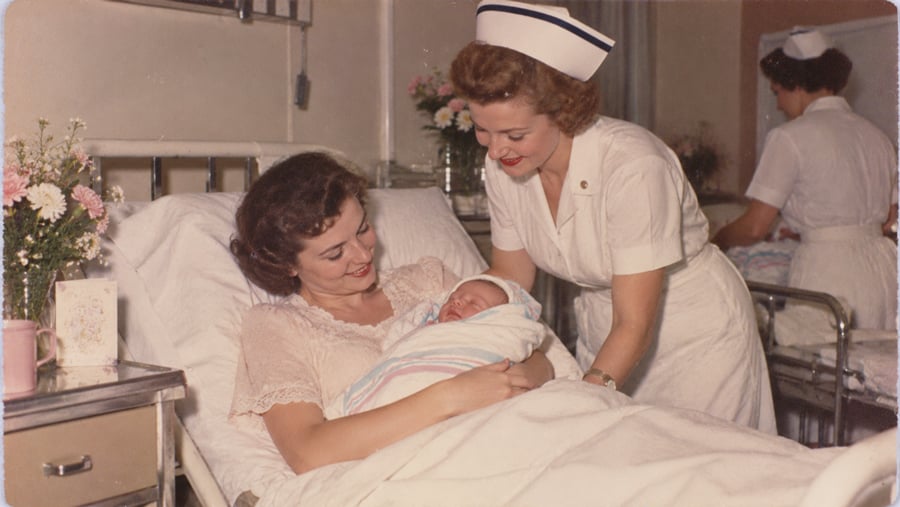 A nurse smiles at a mother holding her newborn baby in a hospital bed; flowers and a card sit on the bedside table, and another nurse is in the background.