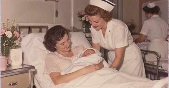 A new mother smiles at her swaddled newborn in a hospital bed while a nurse in a white uniform and cap stands beside them. Another nurse is visible in the background. Flowers and cards are on a nearby table.