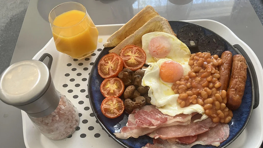 A plate with fried eggs, bacon, sausage, baked beans, mushrooms, grilled tomatoes, black pudding, toast, and a glass of orange juice on a tray with a salt grinder.