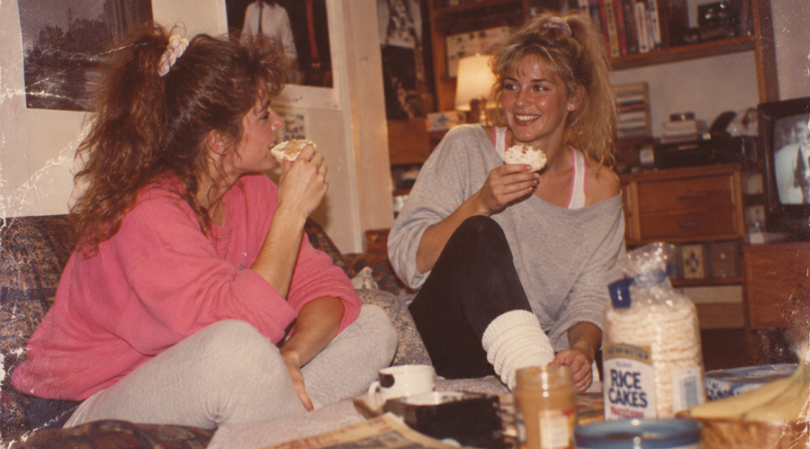 Two women in 1980s-style clothes sit on a couch, smiling and eating rice cakes. A jar of peanut butter and rice cakes package are on the table. The room has posters, books, and a TV in the background.