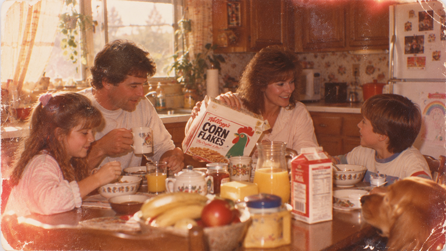 A family of four and a dog eat breakfast at a kitchen table with cereal, fruit, juice, and milk. A woman holds a box of Corn Flakes while everyone smiles and talks together in a cozy, sunlit kitchen.