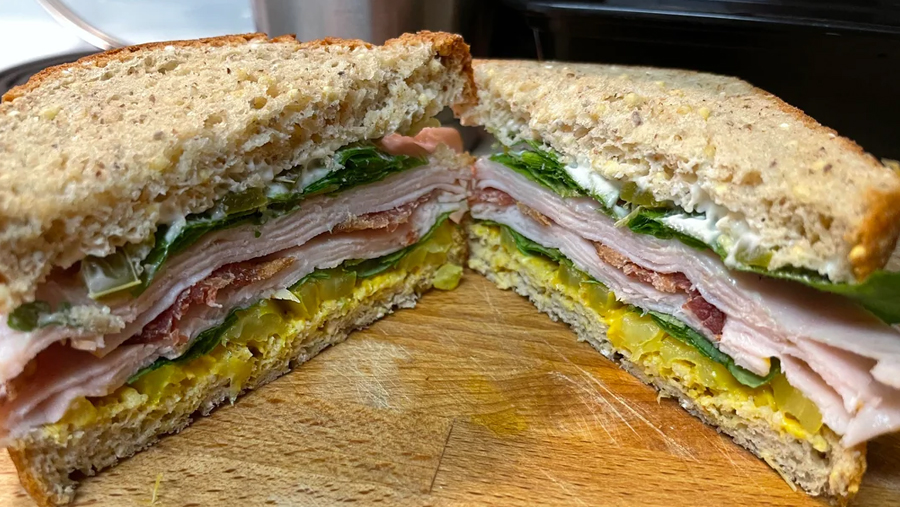 A sandwich cut in half shows layers of turkey, bacon, lettuce, cheese, pickles, and mayonnaise on whole grain bread, set on a wooden cutting board.