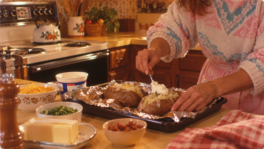 A person in a sweater adds sour cream to baked potatoes on a foil-lined tray in a cozy kitchen, surrounded by toppings like cheese, butter, green onions, and bacon bits.