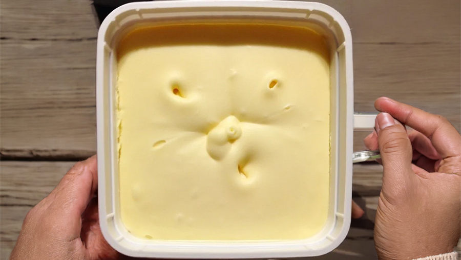 A hand holds a square container of smooth yellow butter with five uneven holes poked in the surface, over a wooden table.