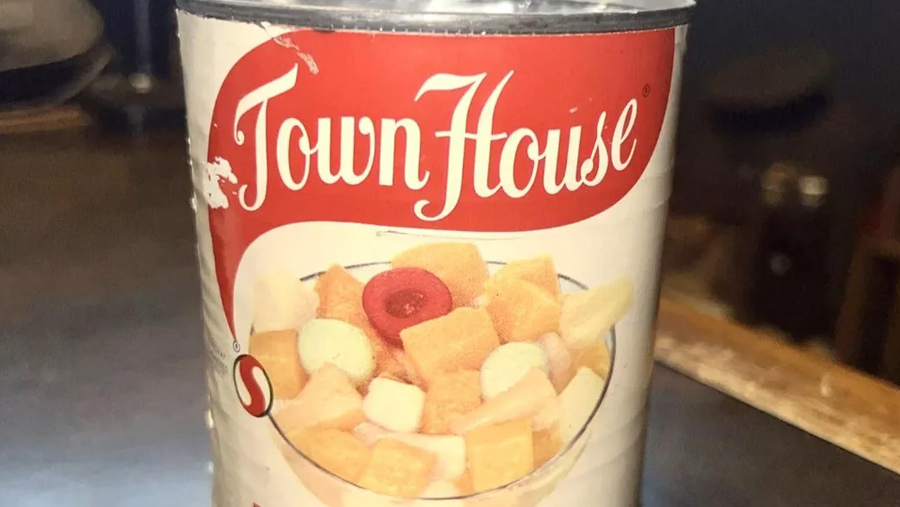 A can labeled “Town House” features an image of a bowl filled with colorful fruit cocktail pieces, including cherries, peaches, and pears. The can sits on a dark surface.