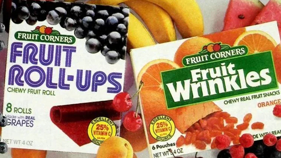 Two boxes of Fruit Corners snacks: Fruit Roll-Ups (grape flavor, left) and Fruit Wrinkles (orange flavor, right), surrounded by various fresh fruits including bananas and cherries.