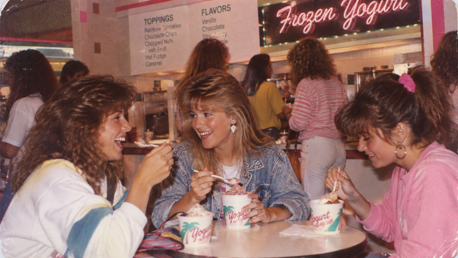 Three women sit at a table in a frozen yogurt shop, laughing and eating yogurt. The background shows a toppings and flavors menu with a pink neon “Frozen Yogurt” sign.