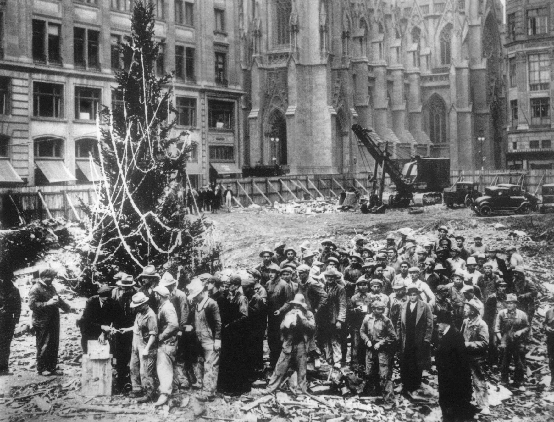 A large group of construction workers in hats stands around a decorated Christmas tree in a dirt lot in front of a church and office buildings.