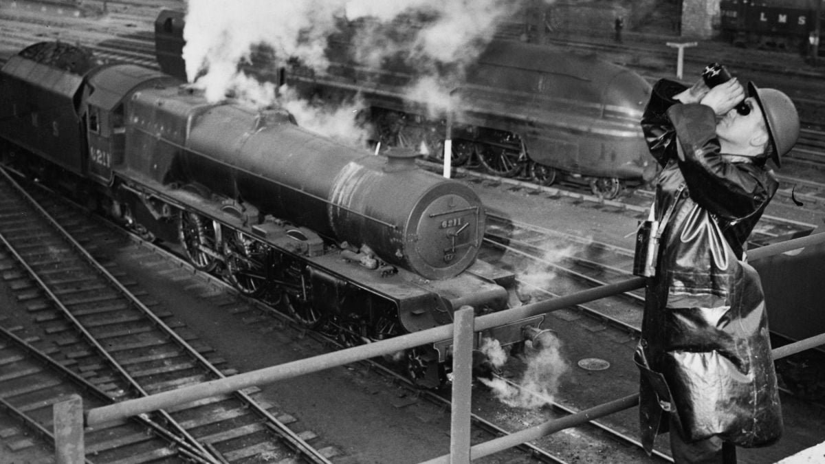 A person in a coat and hat uses binoculars to watch a steam locomotive at a train yard, with rails and other trains visible in the background. Steam rises from the engine.