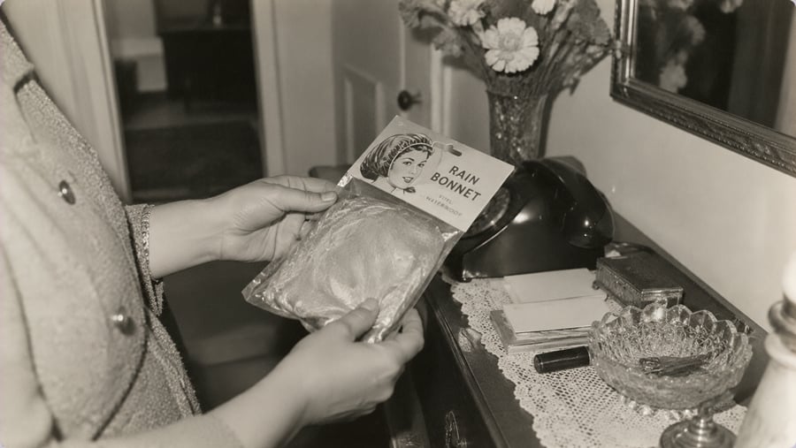 A person holds a packaged rain bonnet in front of a table with a lace cover, a rotary phone, envelopes, a decorative bowl, and a vase of flowers.
