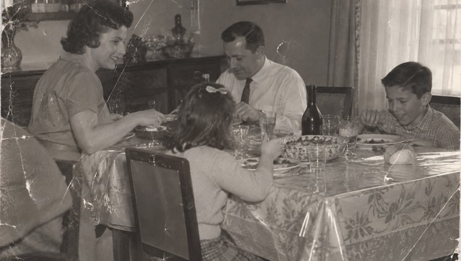A black and white photo of a family of four, two adults and two children, sitting around a dining table and eating a meal together in a cozy, vintage-style room.