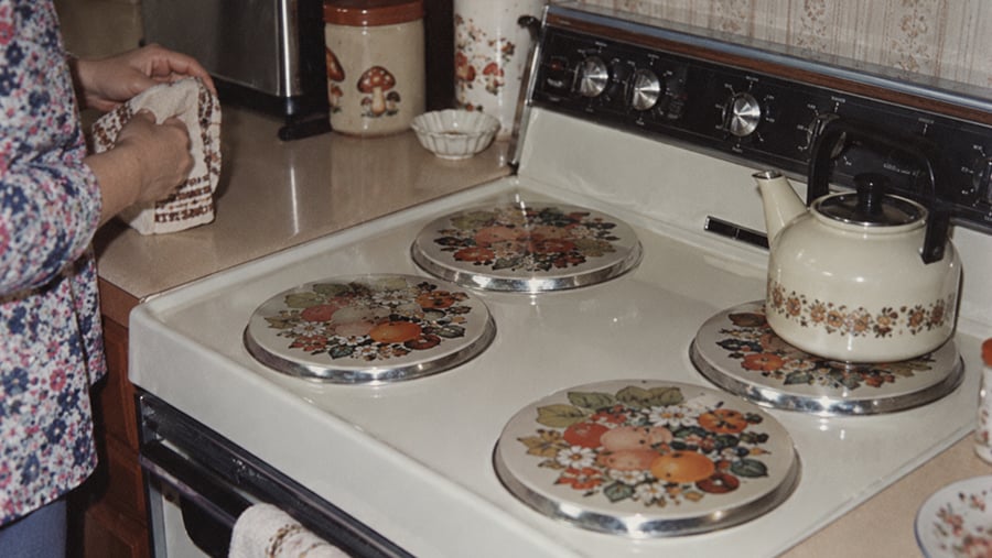 A person stands by a vintage stove with floral burner covers and a floral-patterned kettle. The countertop holds mushroom-themed canisters and a small dish.