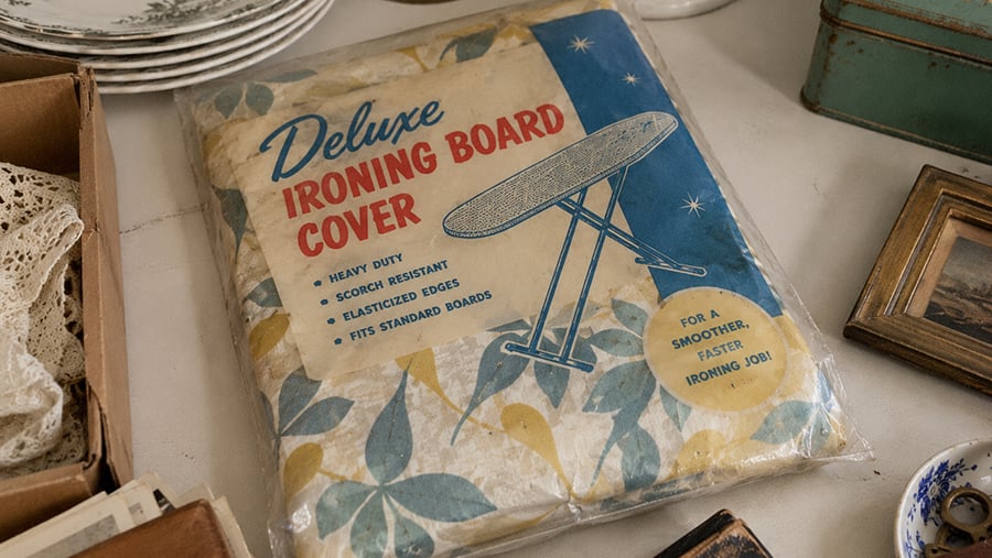 A packaged “Deluxe Ironing Board Cover” with blue and yellow floral print is displayed on a table surrounded by vintage items, including plates and framed pictures.
