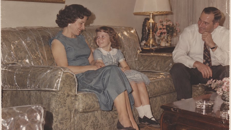 A woman and a girl in blue dresses sit smiling on a sofa with clear plastic cover, while a man in a white shirt and tie sits nearby, all appearing happy in a cozy, vintage living room.