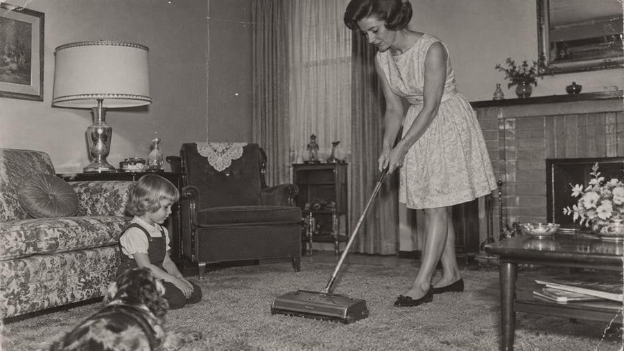 A woman in a dress vacuums a living room carpet while a young girl and a dog watch. The room has a sofa, armchair, lamp, and fireplace, with framed art and curtains in the background.