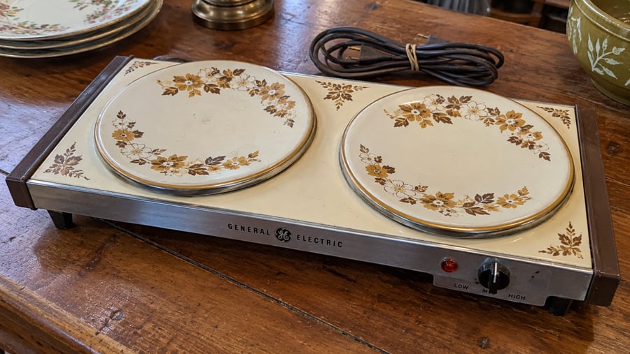 A vintage General Electric double hot plate with floral designs, a dial for low or high settings, and a power cord, sits on a wooden surface.