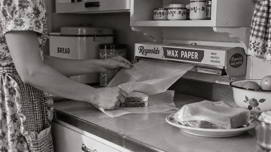 A person in a patterned dress wraps a sandwich in wax paper on a kitchen counter. A bread box, jars, and a plate with another sandwich are nearby. The image is black and white.