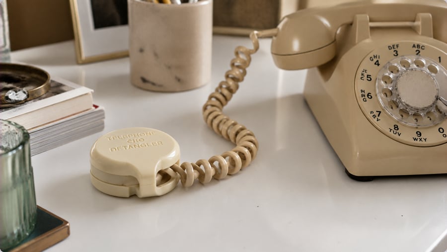 A beige rotary phone sits on a white desk with its cord attached to a round telephone cord detangler. Nearby are a green glass, a book, a tray, and a cup holding pens.