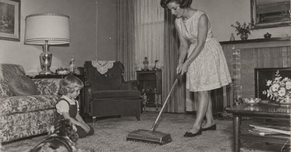 A woman in a dress vacuums a living room carpet while a young girl and a dog watch. The room has vintage furniture, a lamp, and a fireplace.