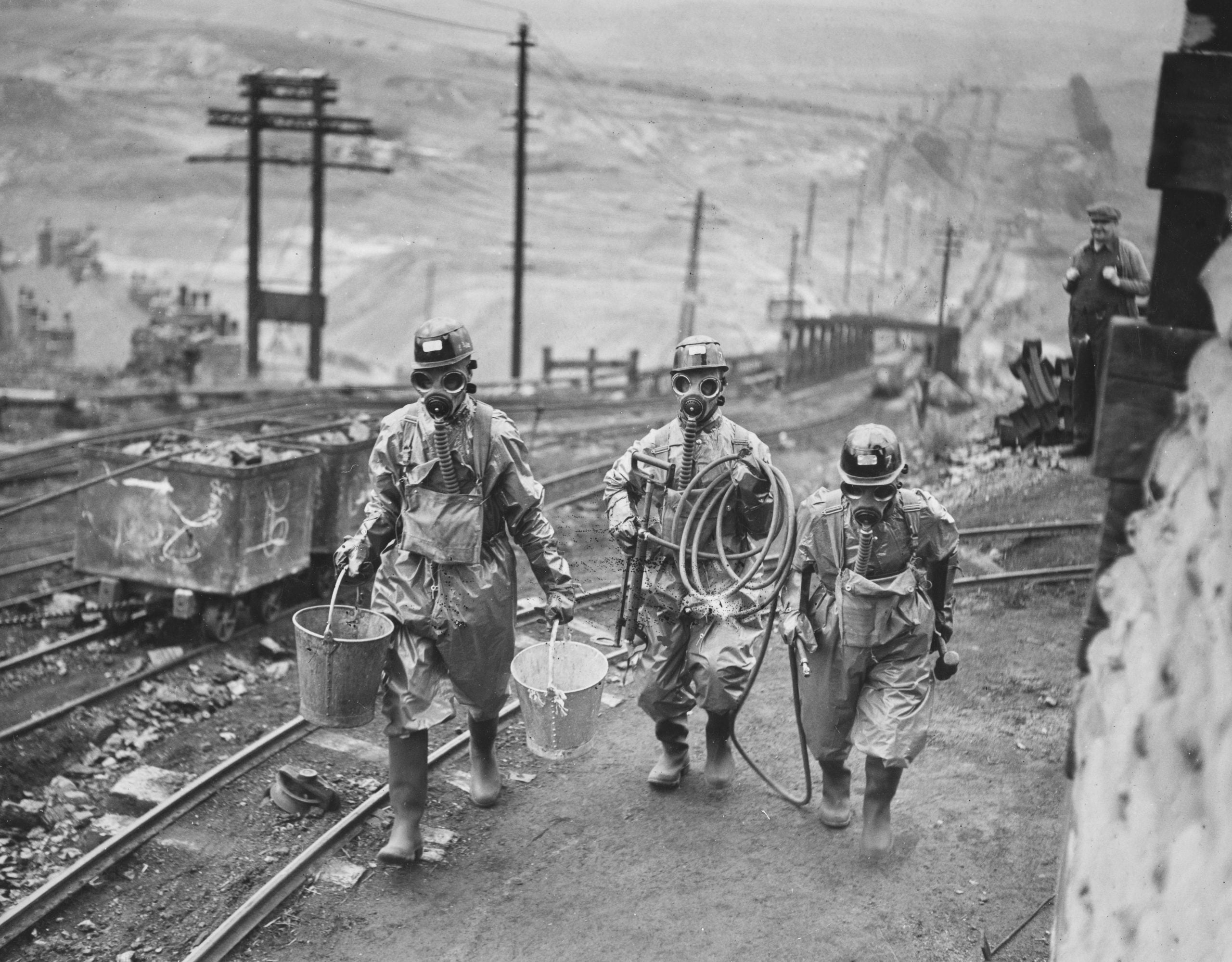 Three people in protective suits and gas masks walk near mining tracks, carrying buckets and equipment. The landscape is industrial, with power poles and distant hills visible in the background.
