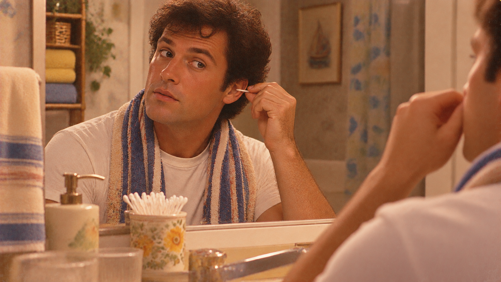 A man wearing a towel around his neck cleans his ear with a cotton swab while looking into a bathroom mirror.