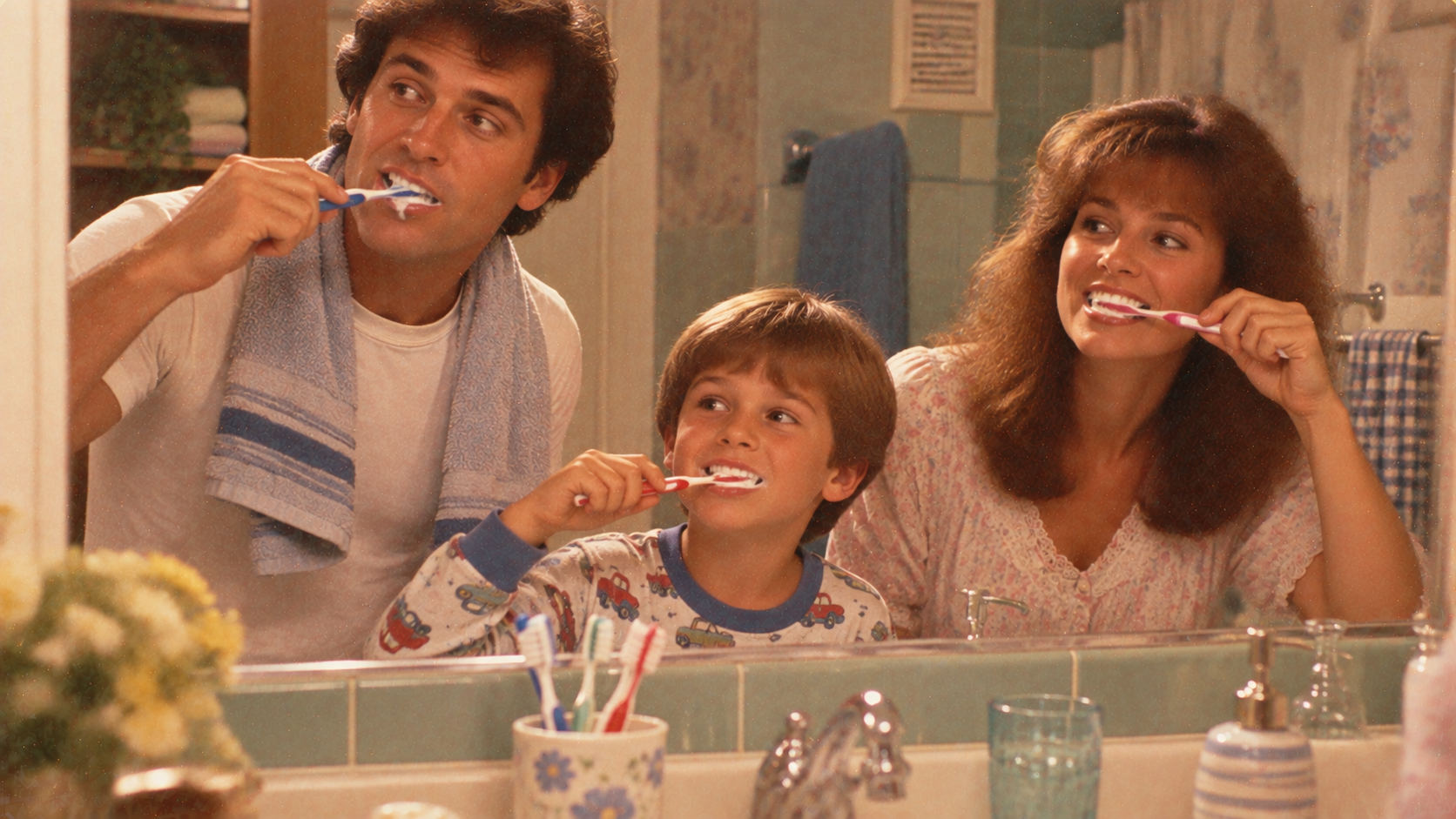 A father, young boy, and mother brushing their teeth together in a bathroom mirror.