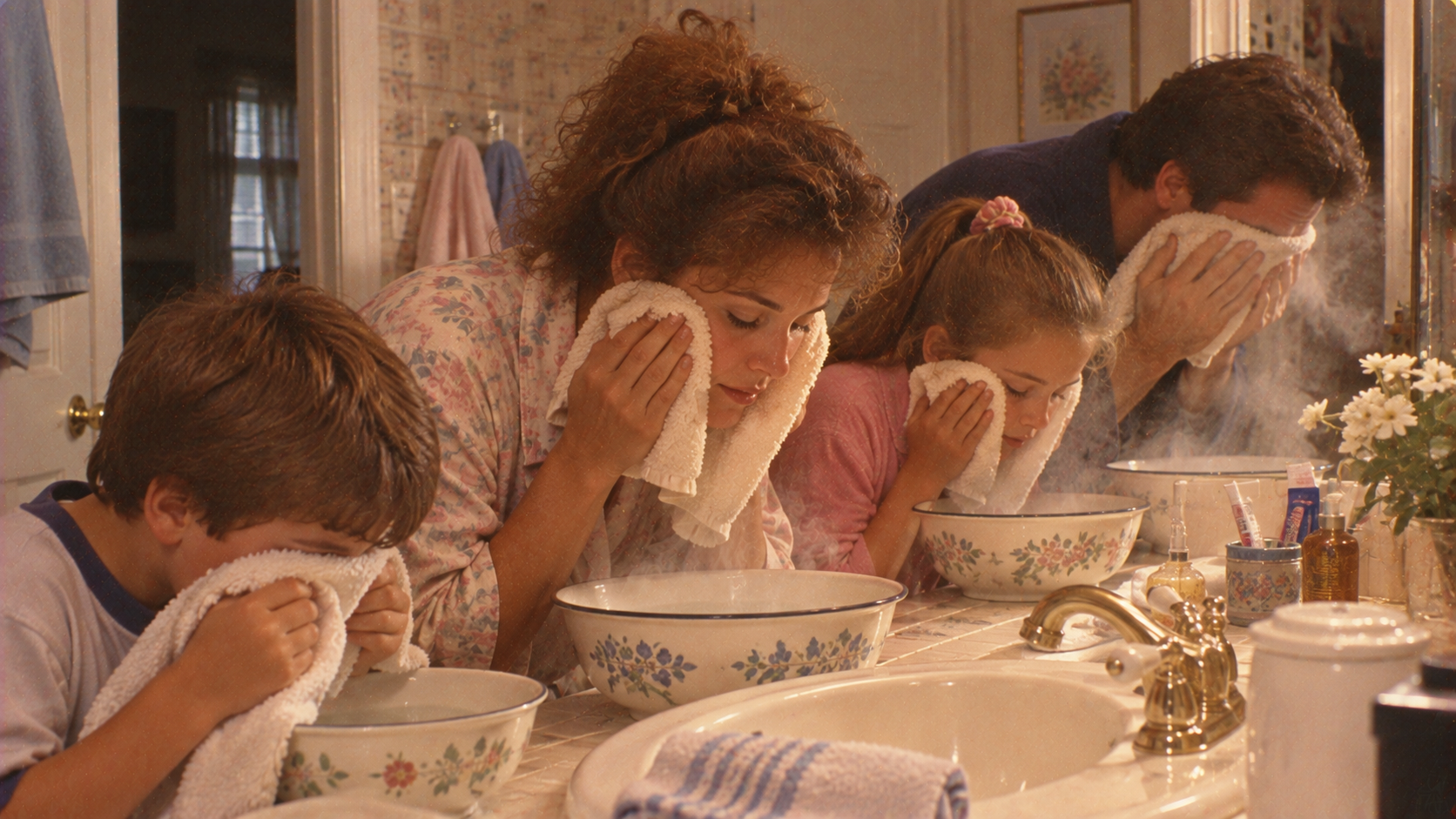 A family of four gathers around a bathroom vanity, each pressing a white washcloth to their face over individual bowls of steaming water.